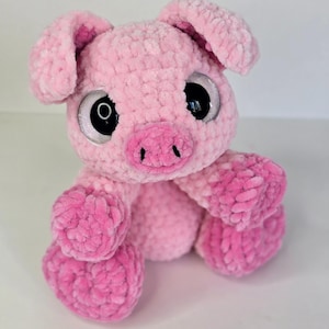 Puede incluir: Un peluche de cerdo rosa hecho a mano con grandes ojos negros brillantes y un hocico rosa. El cerdo tiene orejas ca&iacute;das y est&aacute; sentado erguido, mostrando su cuerpo suave y texturizado. El peluche es de color rosa claro con acentos rosas m&aacute;s oscuros.