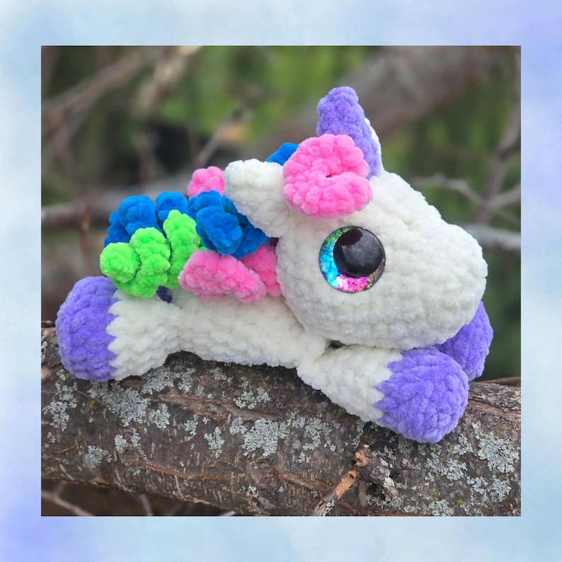 May include: A handmade plush unicorn toy with a white body, purple hooves and horn, and colorful mane and tail accents in pink, blue, and green. The unicorn has large, iridescent eyes and is lying on a tree branch.