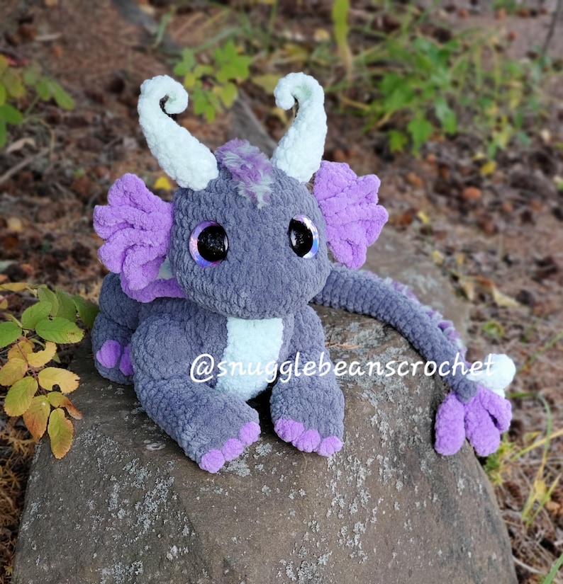 May include: A gray and purple crocheted dragon plush toy with large eyes. The dragon has a long tail and is sitting on a rock. The text "@snugglebeanscrochet" is visible on the dragon's body.