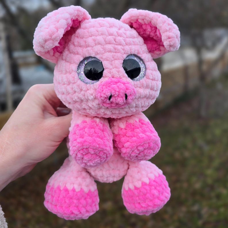 Puede incluir: Un cerdito rosa hecho a mano con ojos grandes y brillantes. El cerdo tiene un cuerpo texturizado, orejas ca&iacute;das y un peque&ntilde;o hocico. Los pies y la parte inferior de las patas son de un tono rosa m&aacute;s oscuro.