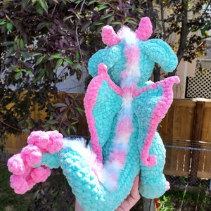 May include: A crocheted plush dragon toy with a light blue and pink colour scheme. The dragon has a fluffy white tail and pink wings.