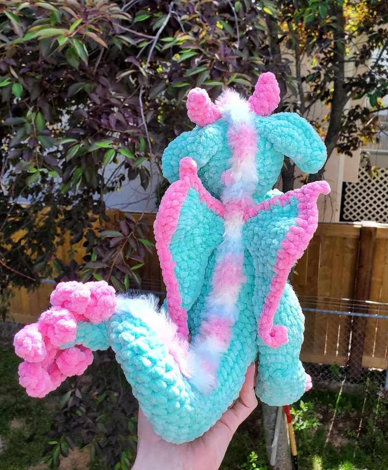 May include: A crocheted plush dragon toy with a light blue and pink colour scheme. The dragon has a fluffy white tail and pink wings.