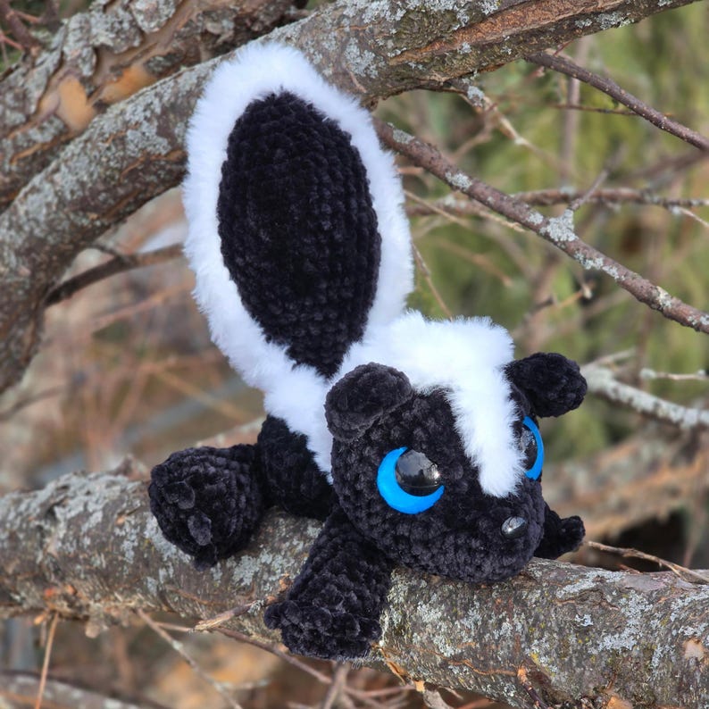 May include: A handmade crochet skunk plush toy with a black body, white stripe, and fluffy tail. The skunk has blue eyes and is posed on a tree branch. The plush toy is a cute, cuddly animal.
