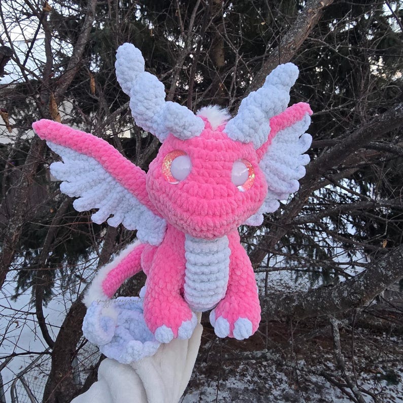 May include: A pink crocheted dragon with white wings and sparkly eyes. The dragon is sitting on a hand.