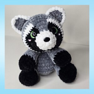 May include: A handmade crochet raccoon plush toy. The plush features a gray and white body with black accents on the paws, eyes, and mask. The raccoon has a black nose and green eyes. The ears are white-trimmed. The plush is sitting upright.