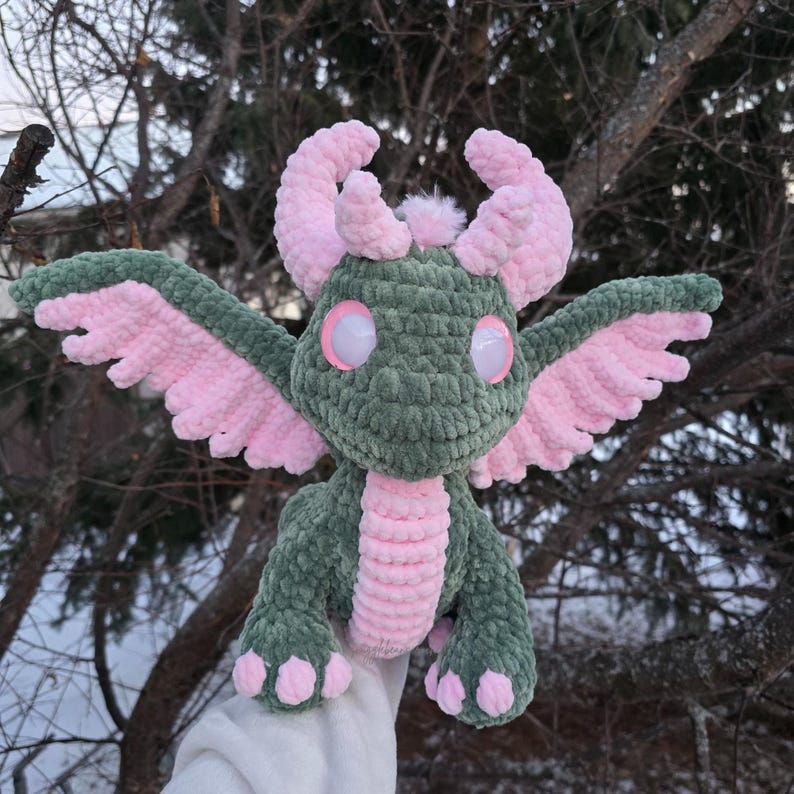 May include: A green and pink crocheted dragon with large white eyes. The dragon has pink wings and a pink tail.