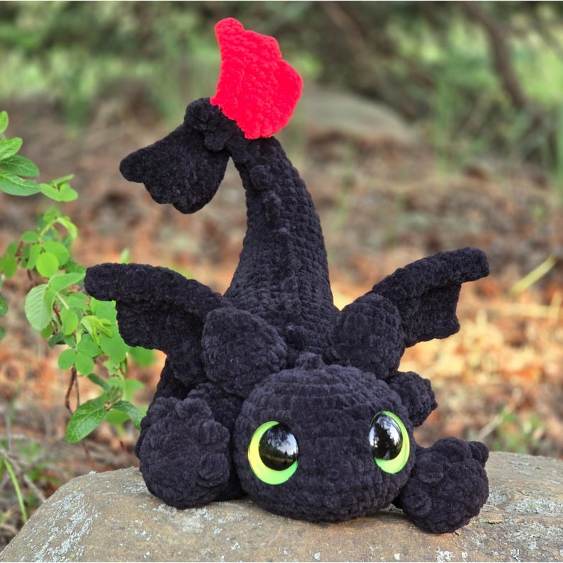 Pode incluir: Um drag&atilde;o de pel&uacute;cia preto de croch&ecirc; feito &agrave; m&atilde;o com uma barbatana vermelha. O drag&atilde;o tem olhos grandes e redondos com &iacute;ris verdes e uma express&atilde;o amig&aacute;vel. A pel&uacute;cia est&aacute; sentada em uma rocha, com um fundo verde desfocado.