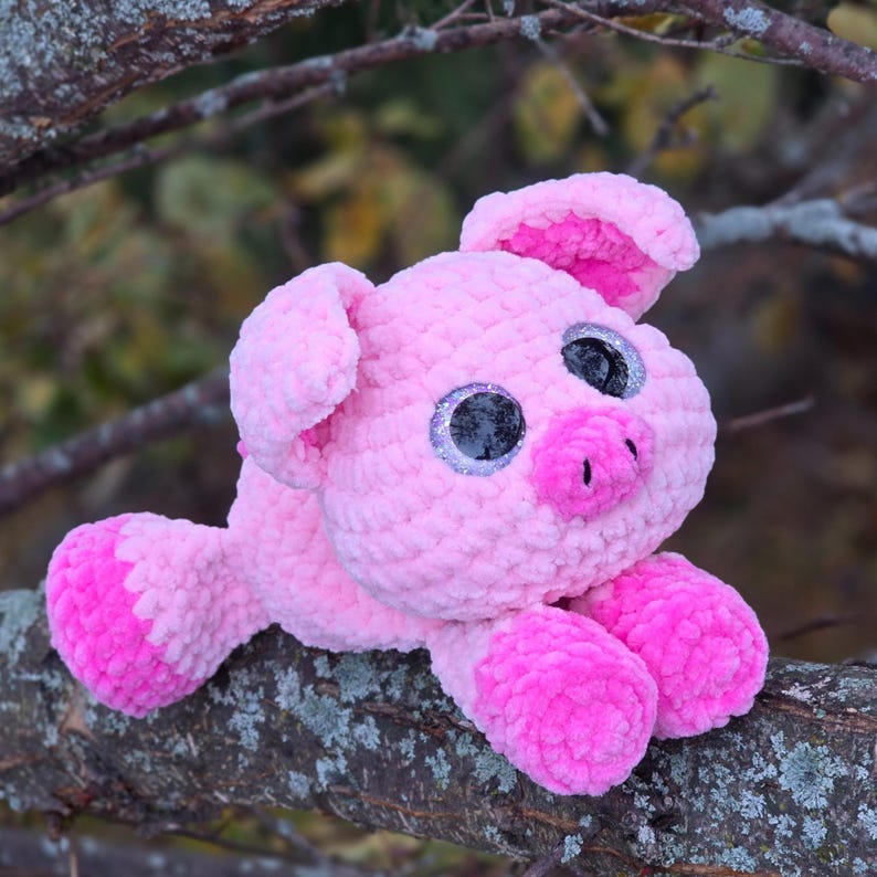 Puede incluir: Un peluche de cerdo rosa hecho a mano con ganchillo, con ojos grandes y brillantes y un hocico peque&ntilde;o. El cerdo tiene detalles en rosa m&aacute;s oscuro en las orejas y las patas. El peluche est&aacute; sobre una rama.