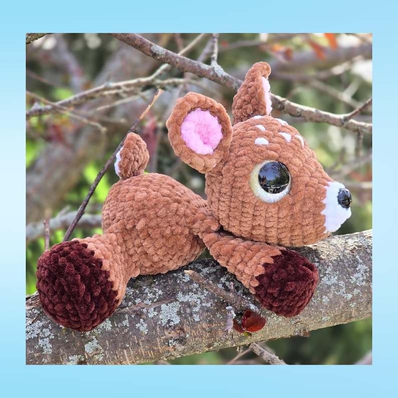 May include: A handmade brown crochet deer plushie with white spots, large black eyes, and pink inner ears. The deer is resting on a tree branch, showcasing its soft, textured body and dark burgundy hooves. A charming woodland creature.
