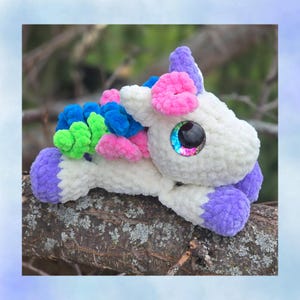May include: A handmade plush unicorn toy with a white body, purple hooves and horn, and colourful mane and tail accents in pink, blue, and green. The unicorn has large, iridescent eyes and is lying on a tree branch.