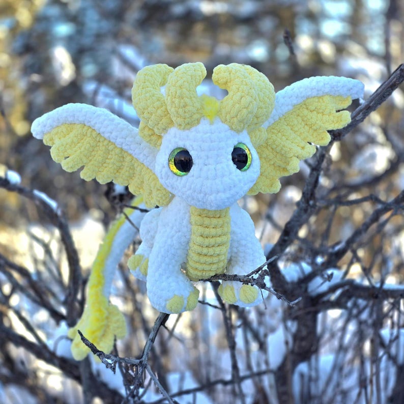 May include: A white crocheted dragon with yellow accents. The dragon has large, green eyes and a long, curled tail. It is sitting on a branch in a snowy woodland.
