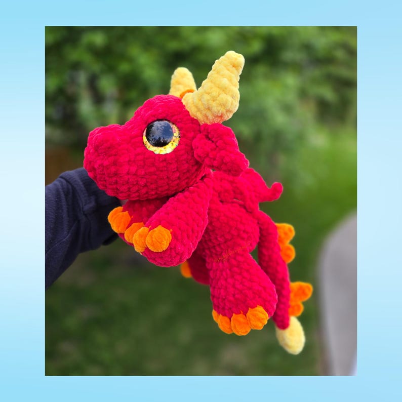 May include: A handmade red crochet dragon with yellow horns and orange accents. The dragon has large, round eyes with black pupils and yellow irises. The plush toy is soft and cuddly, perfect for children or as a decorative item.