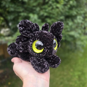 Puede incluir: Un juguete de drag&oacute;n de peluche negro con grandes ojos amarillos brillantes. El drag&oacute;n tiene un aspecto suave y texturizado y est&aacute; acostado. El fondo es verde borroso, lo que sugiere un entorno al aire libre. El objeto est&aacute; hecho a mano.