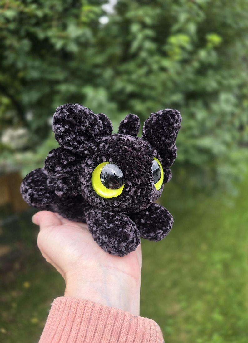 Puede incluir: Un juguete de drag&oacute;n de peluche negro con grandes ojos amarillos brillantes. El drag&oacute;n tiene un aspecto suave y texturizado y est&aacute; acostado. El fondo es verde borroso, lo que sugiere un entorno al aire libre. El objeto est&aacute; hecho a mano.