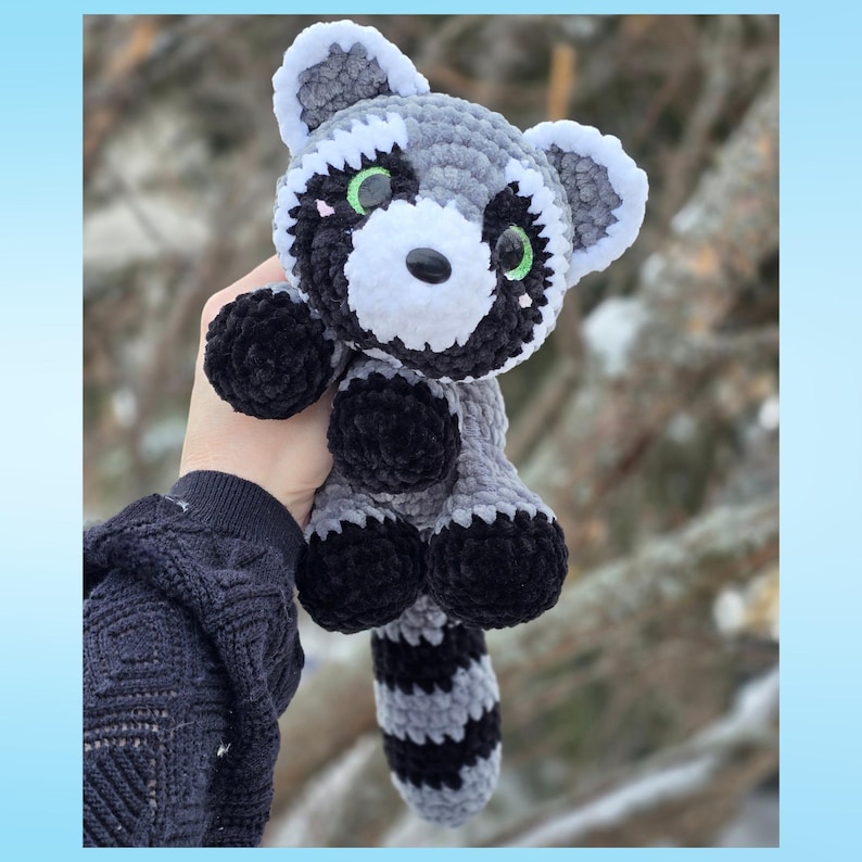 May include: A handmade crochet raccoon plush toy. The stuffed animal features a gray and white body with black accents on the paws, tail, and around the eyes. The raccoon has green eyes and a black nose. The plush is held in a person's hand.