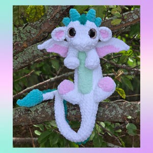 May include: A handmade white plush dragon with large black eyes, pink and white wings, and turquoise accents. The dragon has a mint green belly and pink foot pads. It is perched on a tree branch, showcasing its whimsical design.