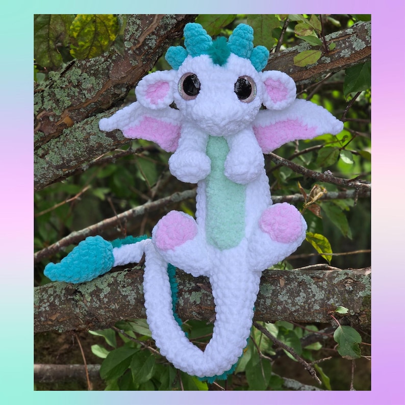 May include: A handmade white plush dragon with large black eyes, pink and white wings, and turquoise accents. The dragon has a mint green belly and pink foot pads. It is perched on a tree branch, showcasing its whimsical design.