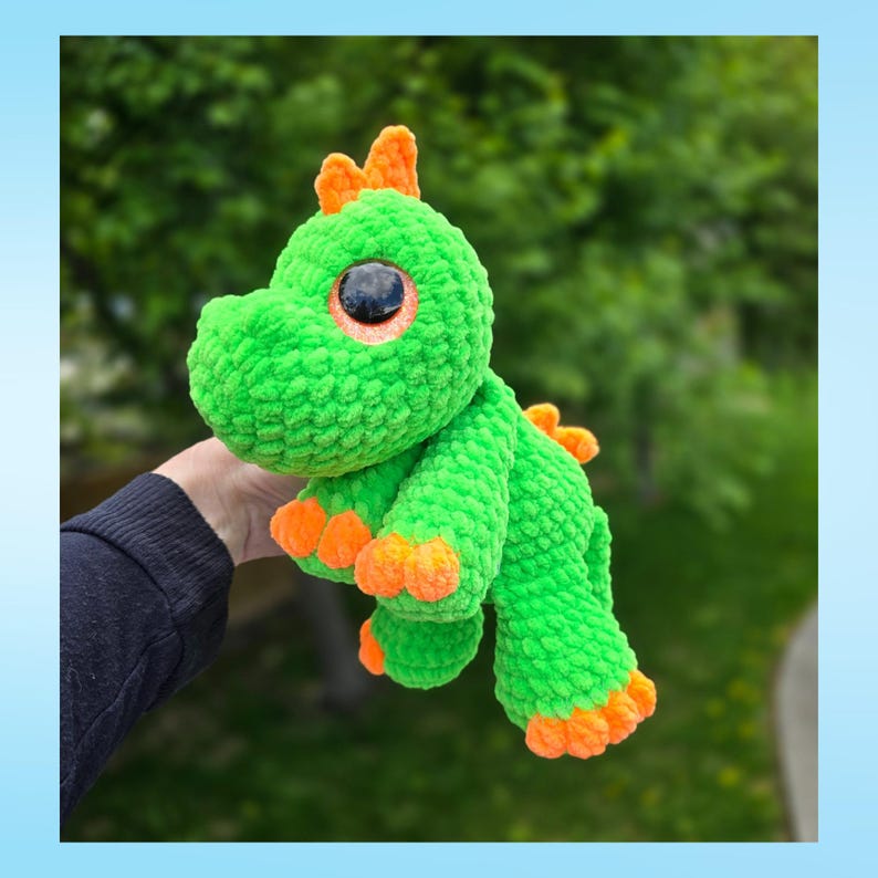 Peut inclure: Un dinosaure vert en crochet fait &agrave; la main avec des d&eacute;tails orange. Le jouet en peluche a de grands yeux ronds et des pointes orange le long du dos. Les pieds du dinosaure et le bout de ses cornes sont &eacute;galement orange. L'arri&egrave;re-plan est une vue floue de feuillage vert.