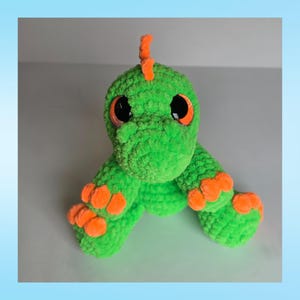 Peut inclure: Une peluche dinosaure verte faite &agrave; la main au crochet, avec des d&eacute;tails orange. Le dinosaure a de grands yeux noirs avec des iris oranges, des griffes oranges et une petite cr&ecirc;te orange. La peluche est en fil doux et textur&eacute;.