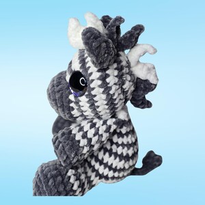 Pode incluir: Um brinquedo de pel&uacute;cia feito &agrave; m&atilde;o em forma de vaca, com um padr&atilde;o xadrez em cinza e branco. A vaca tem olhos pretos grandes e brilhantes e chifres brancos. O brinquedo macio e fofinho est&aacute; sobre um fundo azul claro.