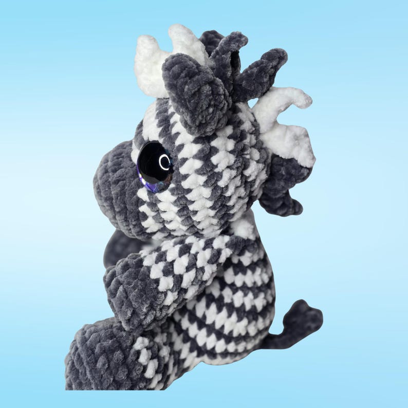 Pode incluir: Um brinquedo de pel&uacute;cia feito &agrave; m&atilde;o em forma de vaca, com um padr&atilde;o xadrez em cinza e branco. A vaca tem olhos pretos grandes e brilhantes e chifres brancos. O brinquedo macio e fofinho est&aacute; sobre um fundo azul claro.