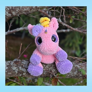 Peut inclure: Une peluche licorne faite &agrave; la main, rose et violette, avec une crini&egrave;re jaune et une petite corne. La licorne a de grands yeux brillants et est assise sur une branche d'arbre. Le jouet est doux et c&acirc;lin, parfait pour les enfants.