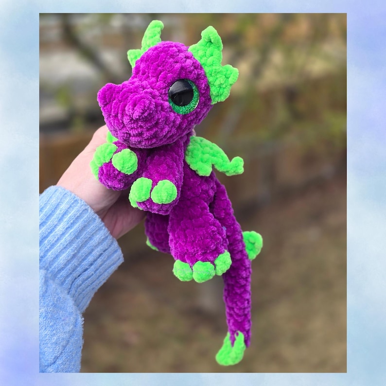 Puede incluir: Un drag&oacute;n de peluche hecho a mano en morado y verde vibrantes. El drag&oacute;n presenta ojos grandes y brillantes, detalles verdes en las alas, pies y cabeza, y una larga cola rizada. El material suave y texturizado le da una apariencia tierna, perfecto para un juguete infantil o un objeto decorativo caprichoso.