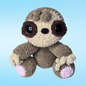 Pode incluir: Um brinquedo de bicho-pregui&ccedil;a feito &agrave; m&atilde;o em croch&ecirc;, em tons de castanho e bege, com olhos grandes e pretos e detalhes em rosa. O brinquedo tem um aspeto macio e texturizado e apresenta um fundo azul claro. As bochechas e os p&eacute;s da pregui&ccedil;a t&ecirc;m detalhes rosa.
