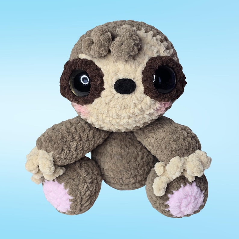 Pode incluir: Um brinquedo de bicho-pregui&ccedil;a feito &agrave; m&atilde;o em croch&ecirc;, em tons de castanho e bege, com olhos grandes e pretos e detalhes em rosa. O brinquedo tem um aspeto macio e texturizado e apresenta um fundo azul claro. As bochechas e os p&eacute;s da pregui&ccedil;a t&ecirc;m detalhes rosa.