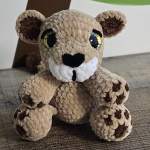 Pode incluir: Um le&atilde;ozinho de pel&uacute;cia bege com detalhes marrons. O animal de pel&uacute;cia feito &agrave; m&atilde;o tem um focinho branco, um nariz preto e olhos grandes e brilhantes. O le&atilde;ozinho est&aacute; sentado, com detalhes marrons nas patas e orelhas.