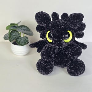 Puede incluir: Un drag&oacute;n de peluche negro hecho a mano con grandes ojos amarillos brillantes. El drag&oacute;n tiene peque&ntilde;as alas y rasgos redondeados. Una peque&ntilde;a planta en maceta con hojas verdes est&aacute; en el fondo. El peluche est&aacute; sobre una superficie blanca.