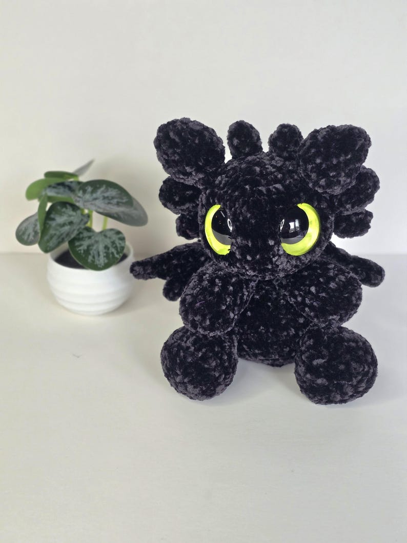 Puede incluir: Un drag&oacute;n de peluche negro hecho a mano con grandes ojos amarillos brillantes. El drag&oacute;n tiene peque&ntilde;as alas y rasgos redondeados. Una peque&ntilde;a planta en maceta con hojas verdes est&aacute; en el fondo. El peluche est&aacute; sobre una superficie blanca.