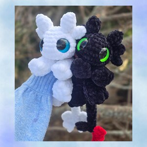 以下が含まれることがあります： 白と黒の2つのぬいぐるみドラゴンが一緒に持たれています。白いドラゴンは青い目を、黒いドラゴンは緑の目を持っています。どちらも小さな翼があり、柔らかく質感のある素材で作られています。黒いドラゴンには赤いアクセントがあります。