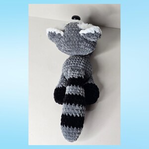 May include: A handmade plush raccoon toy in shades of gray and black. The raccoon has a striped tail, black paws, and white accents on its ears. The toy is made of soft, textured material.