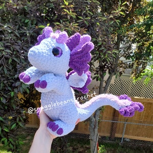 May include: A crocheted purple and light blue dragon plush toy. The dragon has a large head with a single large eye, a long tail, and a small wing on each side. The dragon is being held by a hand.