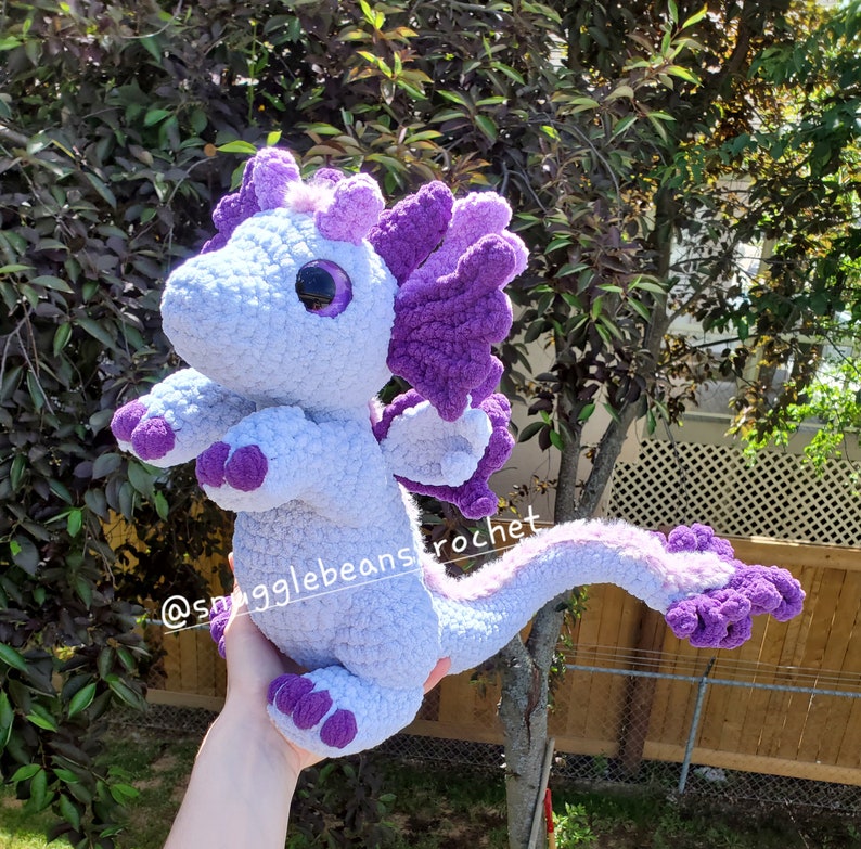 May include: A crocheted purple and light blue dragon plush toy. The dragon has a large head with a single large eye, a long tail, and a small wing on each side. The dragon is being held by a hand.