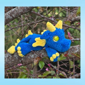Peut inclure: Un jouet de dragon en crochet bleu et jaune fait &agrave; la main, avec de grandes cornes et des accents jaunes. Le dragon a un grand &oelig;il vert et est pos&eacute; sur une branche d'arbre. C'est un jouet doux et en peluche.