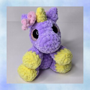 May include: A handmade plush unicorn toy in shades of purple, yellow, and pink. The unicorn has large, black eyes, a yellow horn, and yellow hooves. The plushie is made of soft, textured yarn.