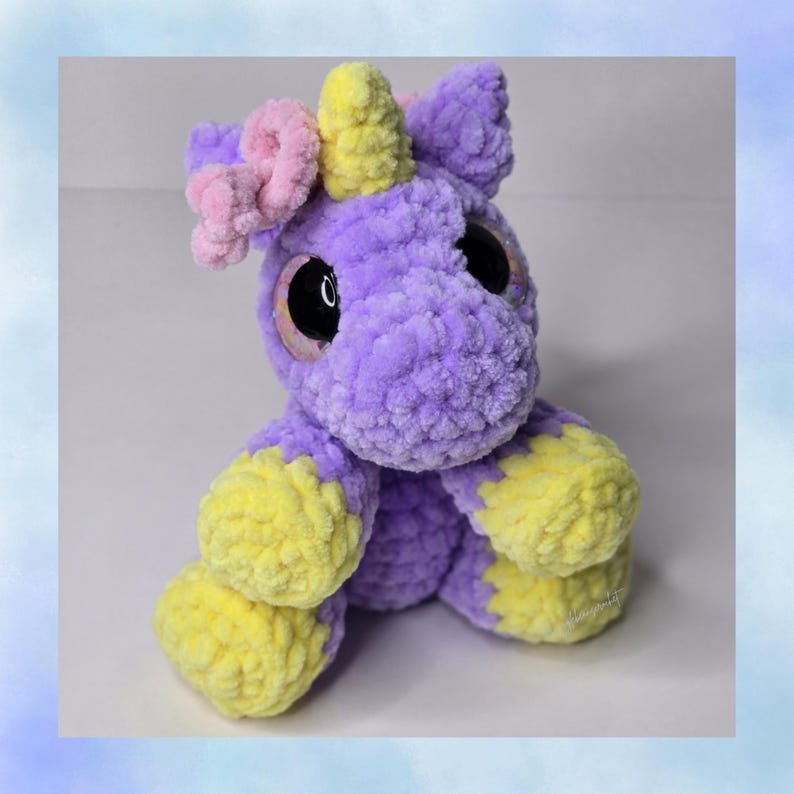 May include: A handmade plush unicorn toy in shades of purple, yellow, and pink. The unicorn has large, black eyes, a yellow horn, and yellow hooves. The plushie is made of soft, textured yarn.