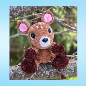 May include: A crocheted deer toy with brown and white spots, large black eyes, and pink inner ears. The deer has a white muzzle and dark brown legs and is sitting on a tree branch. The plushie is a soft, cuddly toy.