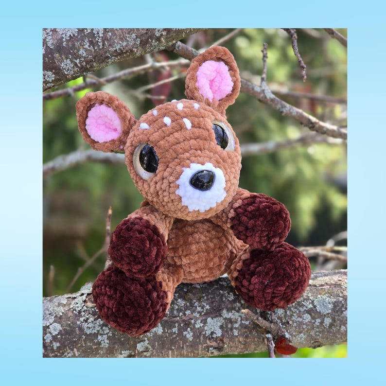 May include: A crocheted deer toy with brown and white spots, large black eyes, and pink inner ears. The deer has a white muzzle and dark brown legs and is sitting on a tree branch. The plushie is a soft, cuddly toy.