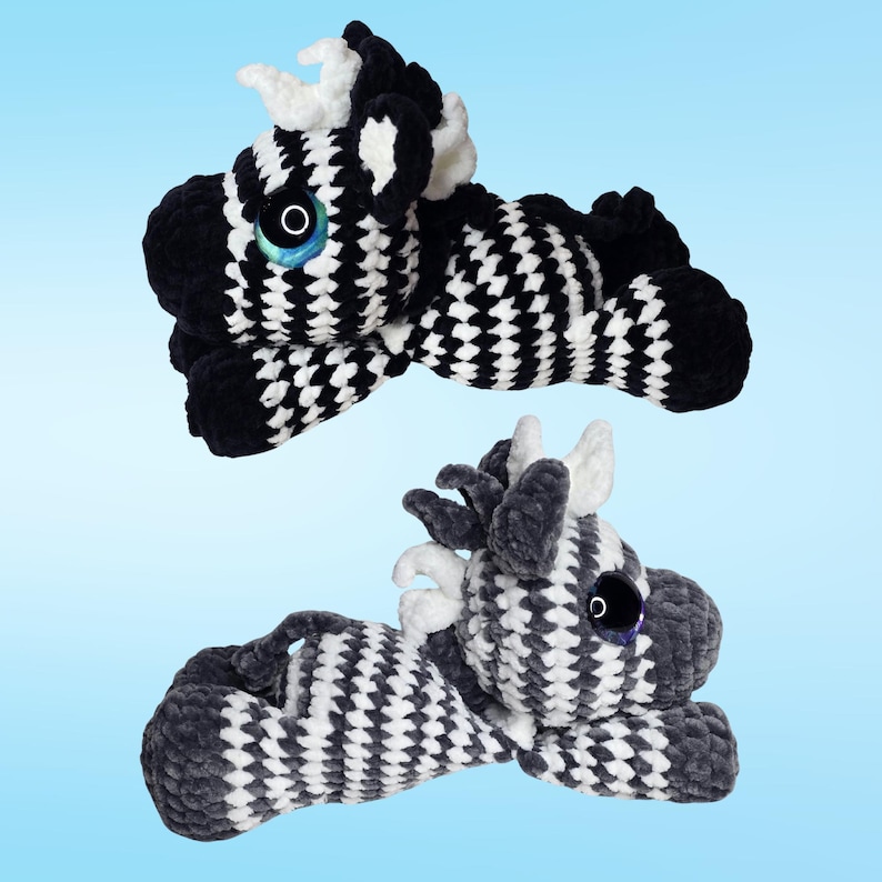 Pode incluir: Dois bichos de pel&uacute;cia de zebra feitos &agrave; m&atilde;o em croch&ecirc;. As zebras est&atilde;o deitadas, com corpos estampados em preto e branco, crinas pretas e chifres brancos. Elas t&ecirc;m olhos grandes e redondos com &iacute;ris azuis. O fundo &eacute; um gradiente azul claro.