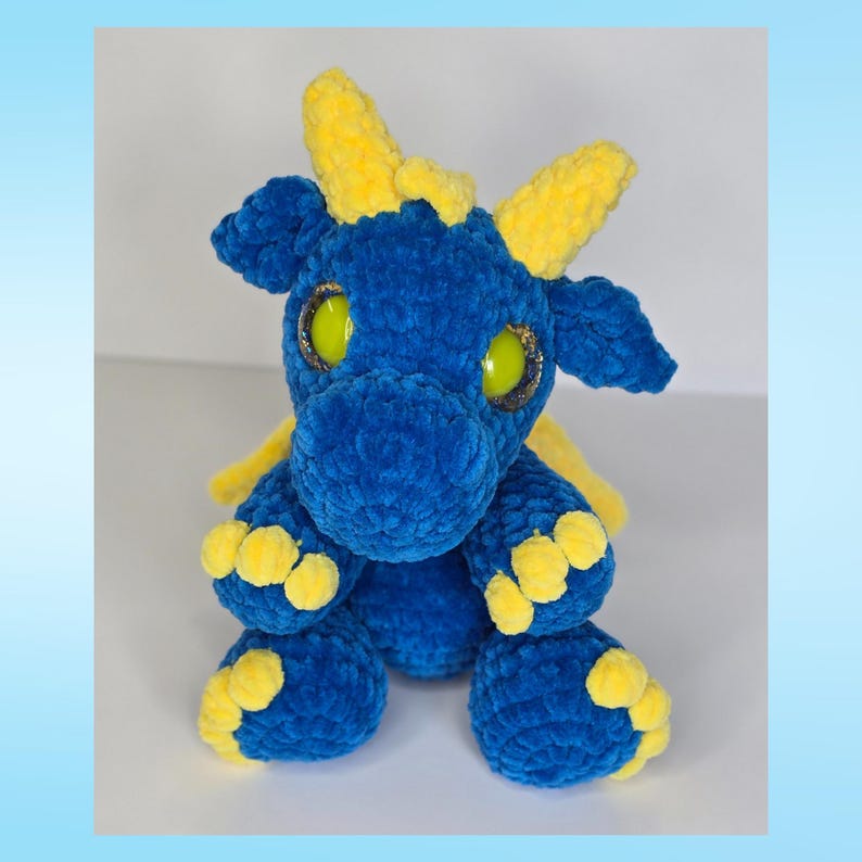 May include: A plush blue dragon with yellow horns, wings, and feet. The dragon has large, round, yellow eyes with black pupils. The dragon is made of a soft, textured material and is sitting upright.