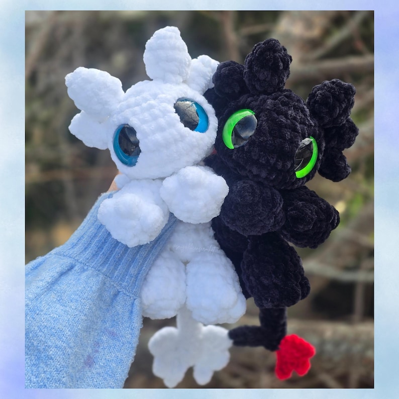 May include: Two crocheted dragon plushies, one white and one black, are held up. The white dragon has blue eyes, and the black dragon has green eyes. Both have small limbs and a tail. The black dragon has a red tail tip.
