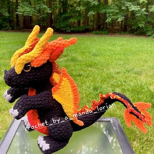 May include: A crocheted dragon toy made with black, orange and yellow yarn. The dragon has a long tail and is standing on a glass table.