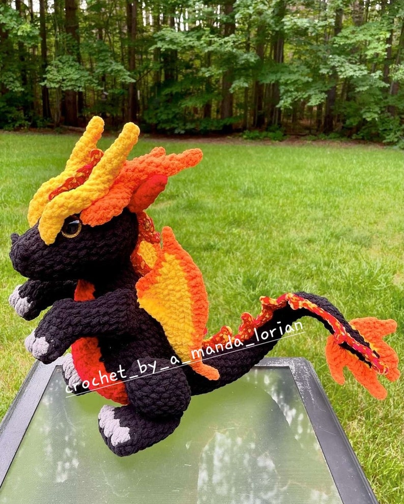 May include: A crocheted dragon toy made with black, orange and yellow yarn. The dragon has a long tail and is standing on a glass table.