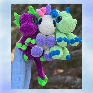 May include: Three crocheted fantasy creatures: a purple dragon, a white unicorn, and a green dragon. The toys have large eyes and are made of soft, textured yarn. The dragon has green accents, and the unicorn has pink and blue accents.