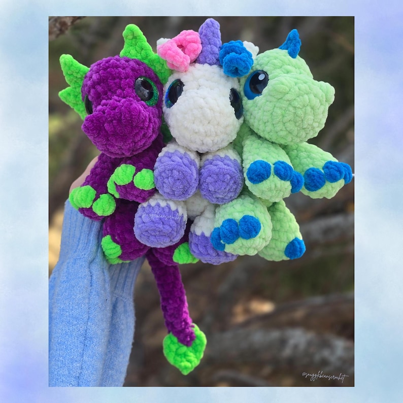 May include: Three crocheted fantasy creatures: a purple dragon, a white unicorn, and a green dragon. The toys have large eyes and are made of soft, textured yarn. The dragon has green accents, and the unicorn has pink and blue accents.