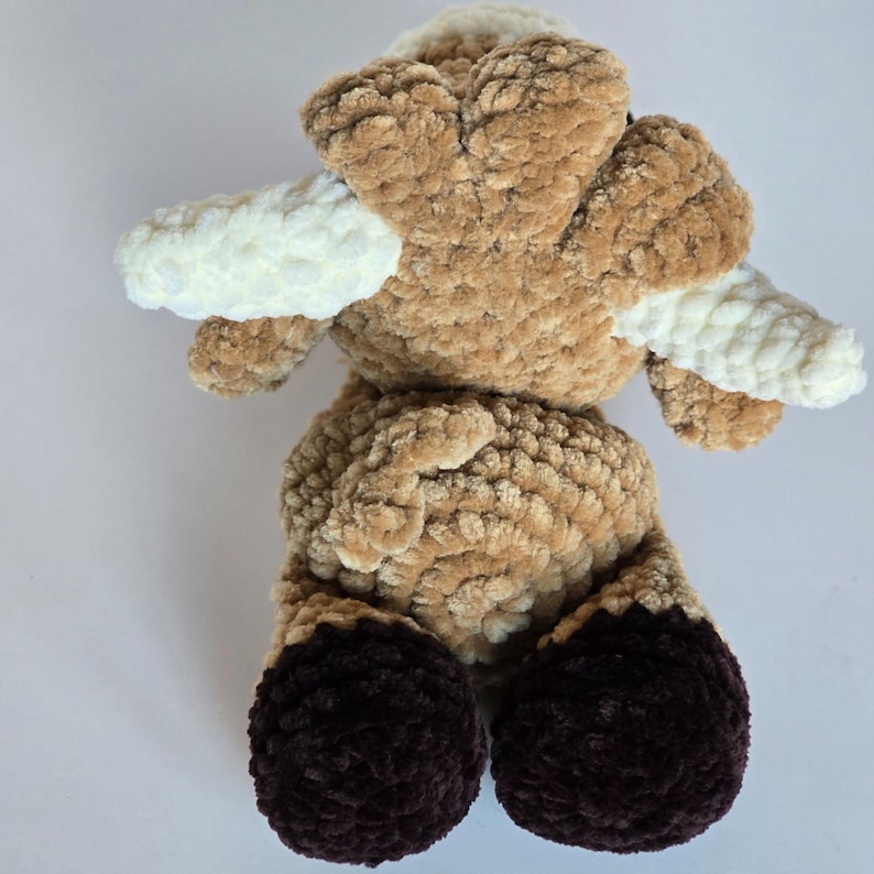 May include: A handcrafted ram-shaped plush toy. It has a soft, textured brown body, white horns, and dark brown hooves. The toy is made from a plush material, giving it a cuddly appearance.