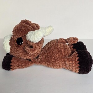 May include: A handmade brown plush bull toy with white horns and large, glittery eyes. The bull is lying down, with dark brown hooves and accents. The plush toy is made of soft, textured yarn.